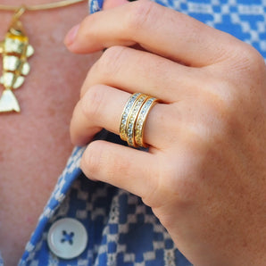 Close-up of a hand wearing gold rings and a bracelet on a blue patterned shirt.